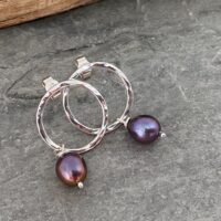 Open circle stud earrings made from hammered silver wire that sparkles when it catches the light. From the bottom of the circle hangs a pretty purple or green small freshwater pearl.