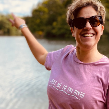 A woman wearing a pink t-shirt with 'Take me to the river' printed across the front while pointing at a river.