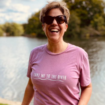 A woman laughing and wearing a pink t-shirt with 'Take me to the river' printed on the front.