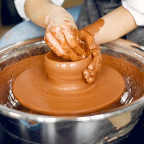 Crop anonymous craftswoman in apron and casual clothes making clay product on pottery wheel  in studio in daylight