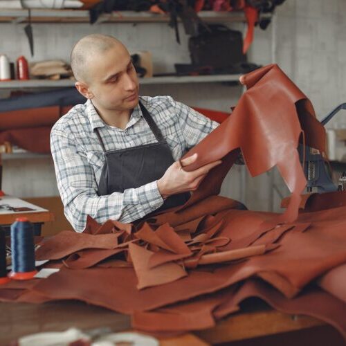 Focused adult male artisan in workwear and apron sitting in workshop among threads and cloths and sewing machines while checking leather material for quality